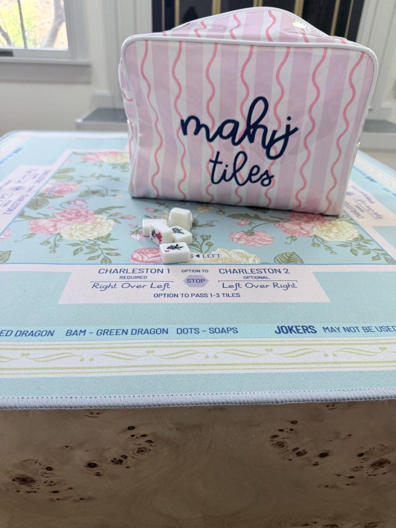 Op de afbeelding: Een roze en witte gestreepte mahjongtegeltas met de woorden "mahj tiles" in marineblauw. De tas staat op een mahjongtafel met bloemenpatroon en tegels. De tafel heeft de tekst "Charleston 1" en "Charleston 2".