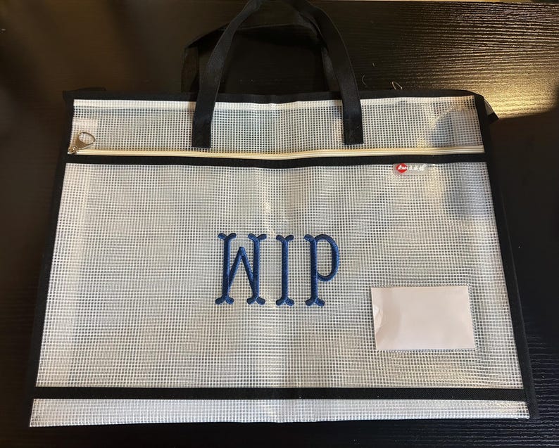 May include: A clear, mesh document bag with black trim and handles. The bag features the embroidered letters "WIP" in blue and a small white label. A zipper runs along the top edge.