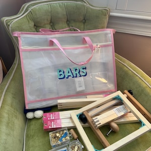 May include: A clear mesh tote bag with pink trim and handles, labeled "BARS" in blue and green lettering, sits on a green armchair. The bag contains craft supplies, including wooden stretcher bars, hardware, and a wooden frame.