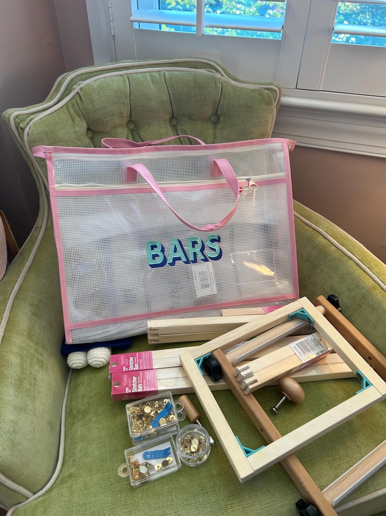 May include: A clear mesh tote bag with pink trim and handles, labeled "BARS" in blue and green lettering, sits on a green armchair. The bag contains craft supplies, including wooden stretcher bars, hardware, and a wooden frame.