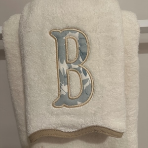May include: A cream-colored hand towel featuring a large embroidered letter B with a blue and white floral pattern inside. The letter is outlined in tan thread, and the towel has a tan hem. Below the letter, the text be our guest is embroidered.