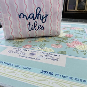Op de afbeelding: Een roze en witte gestreepte Mahjong-tegeltas met de woorden "mahj tiles" in marineblauwe letters. De tas staat op een Mahjong-mat met bloemenpatroon en spelinstructies.