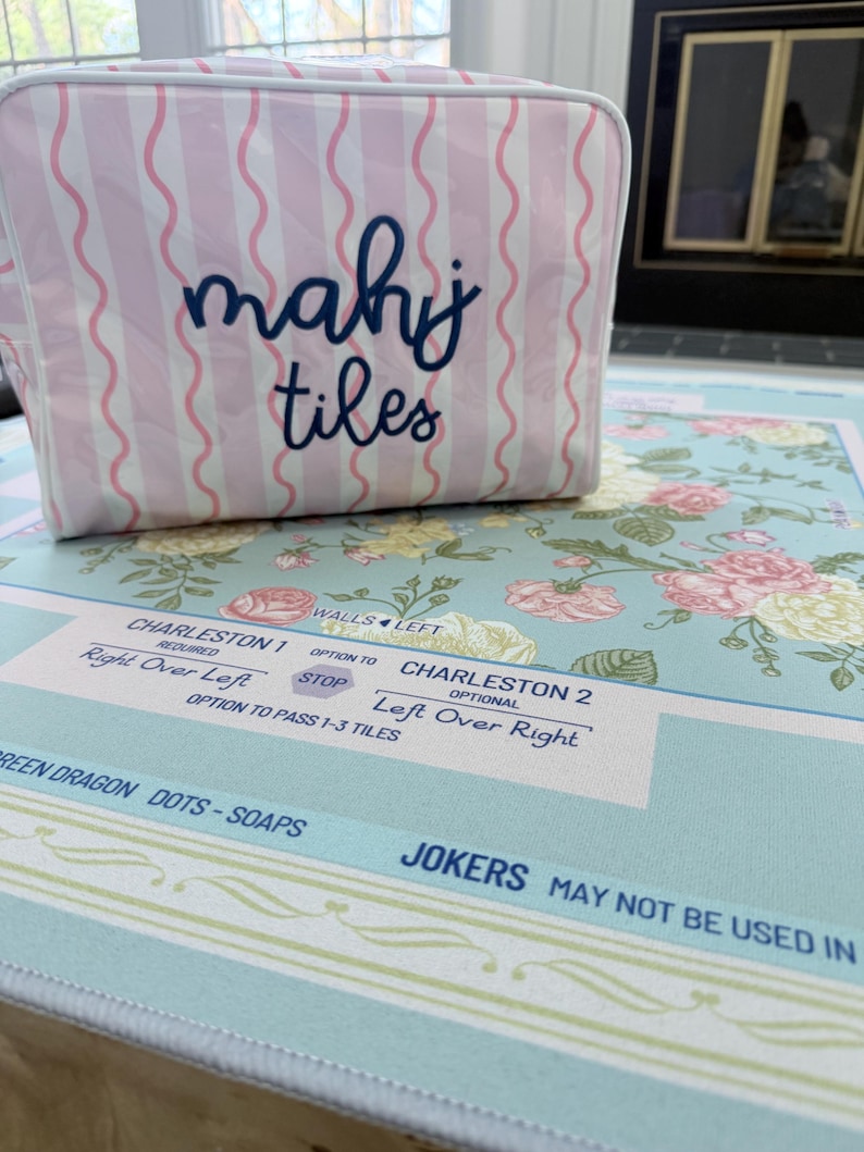Op de afbeelding: Een roze en witte gestreepte Mahjong-tegeltas met de woorden "mahj tiles" in marineblauwe letters. De tas staat op een Mahjong-mat met bloemenpatroon en spelinstructies.