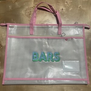 May include: Clear tote bag with pink trim and handles. The bag features a zippered pocket and the word "BARS" embroidered in blue and green. Includes a clear label pocket.