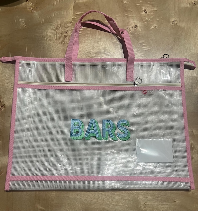 May include: Clear tote bag with pink trim and handles. The bag features a zippered pocket and the word "BARS" embroidered in blue and green. Includes a clear label pocket.
