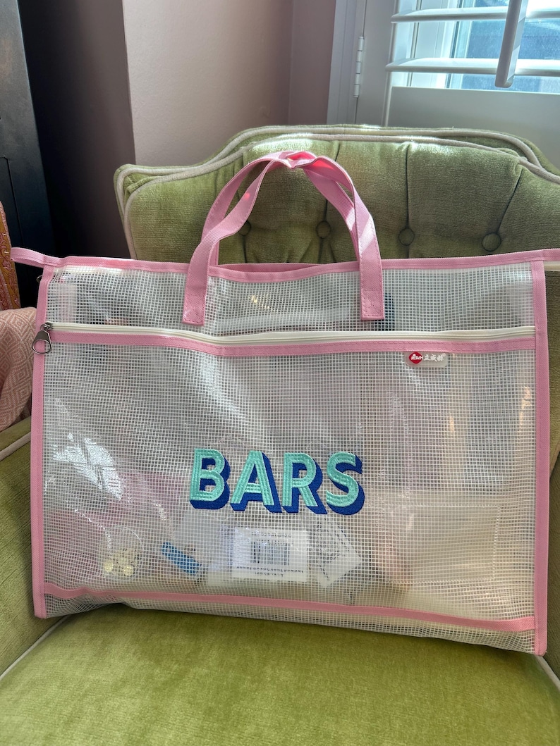 May include: A clear mesh tote bag with pink trim and handles, embroidered with the word "BARS" in teal and blue. The bag is filled with items and sits on a green chair.
