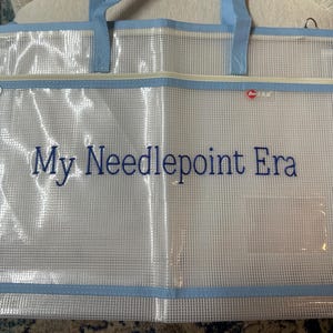 May include: A clear, mesh tote bag with light blue trim and handles. The bag features a zipper closure and the embroidered text "My Needlepoint Era" in blue. A small, red logo is visible.