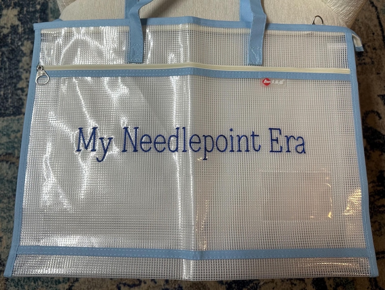 May include: A clear, mesh tote bag with light blue trim and handles. The bag features a zipper closure and the embroidered text "My Needlepoint Era" in blue. A small, red logo is visible.