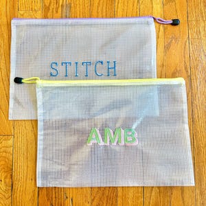 May include: Two clear mesh zippered bags with yellow and purple trim. The top bag has the word "STITCH" embroidered in blue thread. The bottom bag has the word "AMB" embroidered in green thread.