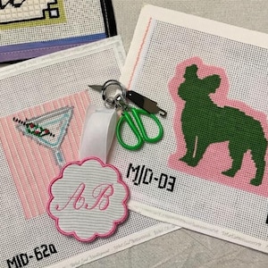 May include: Needlepoint canvases featuring a martini, a green dog silhouette, and a floral-shaped tag with the initials "AB". The canvases are on a white background with pink accents. A pair of green scissors and a key ring are also present.