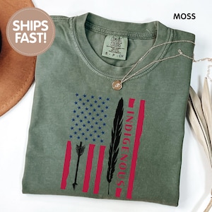 May include: Moss green t-shirt featuring a design with a flag-like graphic. The graphic includes blue stars, red stripes, an arrow, a black feather, and the word "INDIGENOUS" in red. A gold necklace rests on the shirt.