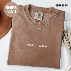 以下が含まれることがあります： エスプレッソブラウンの半袖クルーネックTシャツ。胸元には「caution: may bite」という白い文字がプリントされています。襟元近くの生地の上には、小さな円形のペンダントが付いたシルバーカラーのネックレスが置かれています。
