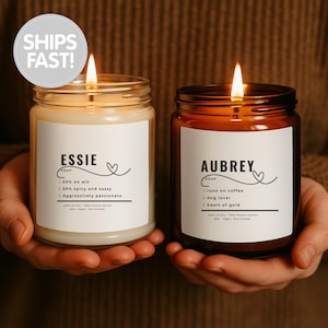 Puede incluir: Dos velas encendidas en frascos de vidrio con etiquetas blancas. Una vela es de color claro, la otra es oscura. Las etiquetas dicen "ESSIE" y "AUBREY" con descripciones debajo. La imagen incluye el texto "SHIPS FAST!"