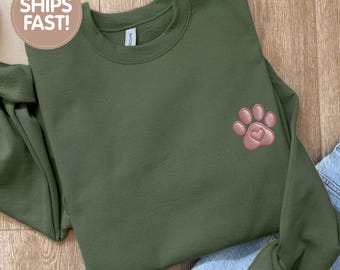 May include: Military green sweatshirt with a pink paw print and heart design. The sweatshirt is made of a soft material. The text "MILITARY GREEN" is printed on the top right of the sweatshirt.