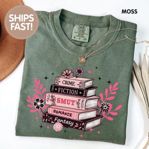 May include: Moss green t-shirt with a stack of books featuring the words "Crime", "Fiction", "Smut", "Romance", and "Fantasy" in a pink and black design. The shirt has a crew neck and short sleeves. A gold necklace is draped across the shirt.