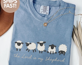 May include: A blue jean colored short-sleeved t-shirt featuring a graphic of five white and black sheep in a row. Below the sheep, the text reads, the Lord is my Shepherd. The shirt has a crew neckline and a Comfort Colors brand label inside.