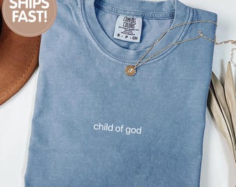 May include: A blue jean colored t-shirt with a crew neckline and the text child of god embroidered in white on the chest. The shirt features a Comfort Colors brand label at the inner neck and is paired with a thin gold chain necklace with a round pendant.