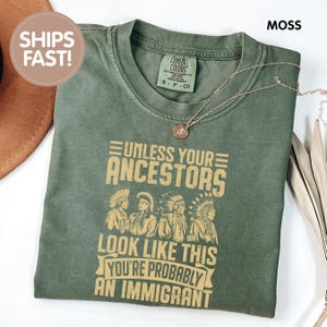 May include: A moss green t-shirt with the text "UNLESS YOUR ANCESTORS LOOK LIKE THIS YOU'RE PROBABLY AN IMMIGRANT" in a tan color. The shirt also features an illustration of four Native American figures. A gold necklace is draped across the shirt.