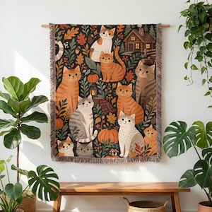 May include: A woven wall tapestry featuring a dark background with multiple cats, pumpkins, autumn leaves, and a small wooden cabin. The cats are depicted in various shades of orange, gray, and white, interspersed with foliage and forest elements.