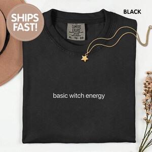 Op de afbeelding: Een zwart t-shirt met de witte tekst "basic witch energy" op de borst gedrukt. Het shirt heeft een ronde hals en een Comfort Colors merklabel aan de binnenkant van de kraag. Een goudkleurige ketting met een kleine sterhanger ligt er bovenop.