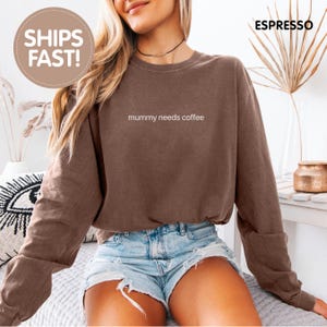 Könnte beinhalten: Ein braunes Langarm-Sweatshirt mit Rundhalsausschnitt und dem weißen Schriftzug "mummy needs coffee" auf der Brust. Das Kleidungsstück hat eine lockere Passform mit gerippten Bündchen am Ärmel und am Halsausschnitt in einem einheitlichen Braunton.