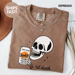 May include: Espresso-colored t-shirt featuring a white skull holding a Sunkist soda can with the text "ZERO SUGAR". The phrase "'til death" is printed below the skull. A silver necklace with a pendant is also visible.