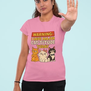 May include: Pink t-shirt with the text "WARNING: I HAVE MAJOR CAT-TITUDE TODAY" above a graphic of three cartoon cats. The cats are orange, white, and black, each with a unique expression. The shirt is being worn by a person.