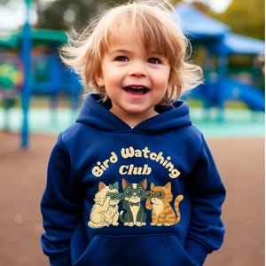 Puede incluir: Una sudadera con capucha azul marino con el texto "Bird Watching Club" y tres gatos de dibujos animados con gafas. Los gatos son blancos, negros y naranjas. El niño sonríe.