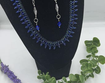 May include: A blue beaded necklace with a scalloped edge and matching earrings with a pearl and crystal accents. The necklace and earrings are displayed on a black velvet stand.