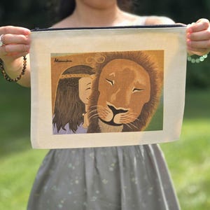 May include: A beige canvas zipper pouch featuring a colorful illustration of a lion and a woman. The lion has a golden mane and closed eyes, and the woman has long brown hair. The pouch is held up in front of a blurred green background.