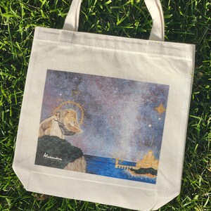 May include: Cream-colored tote bag with a colorful illustration. The artwork depicts a lion with a halo, overlooking a seascape with a city and stars. The bag has two handles and the artist's signature.