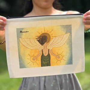 May include: A canvas zipper pouch featuring an illustration of an angel with outstretched wings, a halo, and two suns. The angel is depicted in a green dress against a yellow and blue background. The artist's signature, "Alexandra," is in the upper left corner.