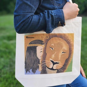 Op de afbeelding: Een witte draagtas met een kleurrijke illustratie van een leeuw en een vrouw. De leeuw heeft een bruine manen en een zwarte neus. De vrouw heeft lang zwart haar en draagt een kroon. De naam "Alexandra" is cursief geschreven.