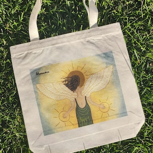 May include: Cream-colored tote bag with a painting of an angel with outstretched wings and a halo. The angel is wearing a green dress and holding two golden orbs. The bag has cream-colored handles and is resting on green grass.