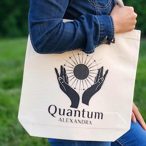 May include: Cream-colored tote bag with a black graphic of a sunburst design held by two hands. The words "Quantum" and "ALEXANDRA" are printed below the graphic. The bag is being held by a person wearing a denim jacket.