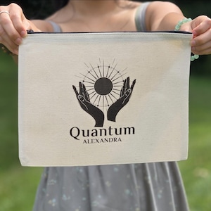 May include: Cream-colored canvas pouch with a black zipper, featuring a black graphic of hands cradling a sunburst design. The text "Quantum ALEXANDRA" is printed below the graphic. The pouch is held up in front of a blurred green background.