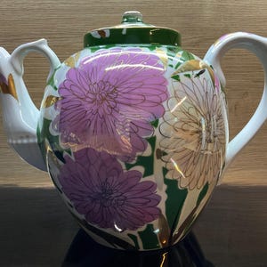 Könnte beinhalten: Eine weiße Keramik-Teekanne mit floralem Design. Die Teekanne zeigt große lila und cremefarbene Blumen mit goldenen Akzenten auf einem grünen und weißen Hintergrund. Der Deckel und der Griff sind weiß.