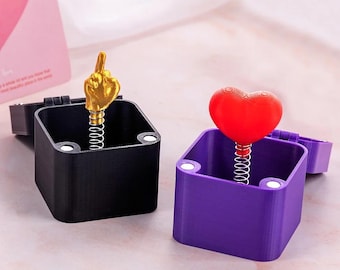 May include: Two novelty boxes, one black and one purple, each with a lid and a spring-loaded surprise. The black box features a gold hand giving the middle finger, while the purple box has a red heart.