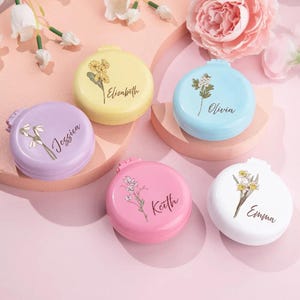 Peut inclure: Plusieurs miroirs compacts personnalisés aux couleurs pastel : lavande, jaune, bleu, rose et blanc. Chaque miroir présente un motif floral et un nom : Jessica, Elizabeth, Olivia, Keith et Emma. Les miroirs sont disposés sur une surface rose.