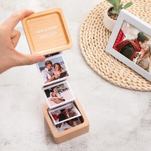 以下が含まれることがあります： 木製のフォトボックス。スライド機構で一連の写真が展開されます。「Jack & Millie」と日付「2024.11.05」が刻印された蓋。白いフォトフレームも表示されています。