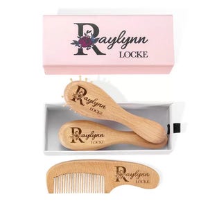 Puede incluir: Un juego de cuidado del cabello de madera, que incluye un cepillo y un peine, presentado en una caja rosa. El juego presenta el nombre "Raylynn Locke" con detalles florales. El cepillo tiene cerdas, mientras que el peine tiene dientes finos.