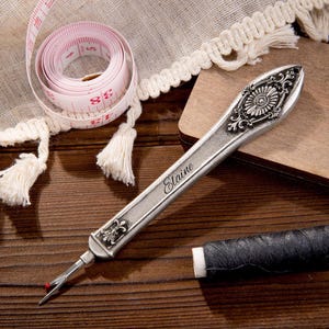 May include: A silver seam ripper with floral detailing and the name "Elaine" engraved on the handle. A pink and white measuring tape and black thread spool are also visible, suggesting a sewing or crafting theme.