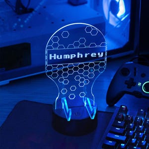 May include: A blue LED lamp with the name "Humphrey" illuminated in white. The lamp has a honeycomb design and sits on a black base. A keyboard and game controller are visible in the background.