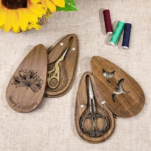 Könnte beinhalten: Holzschereetuis mit floralen und Vogelmotiven. Die Etuis sind geöffnet und zeigen goldene und silberne Scheren. Der Text "Mary's scissors" ist auf den Etuis eingraviert. Garnrollen in Burgunderrot, Grün und Marineblau sind ebenfalls vorhanden.