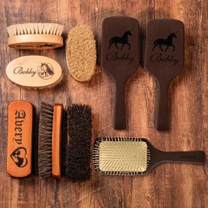 May include: A collection of horse grooming brushes in various shapes and sizes, including oval, rectangular, and paddle styles. Some brushes are engraved with names and unicorn designs. The brushes are made of wood and bristles, arranged on a wooden surface.