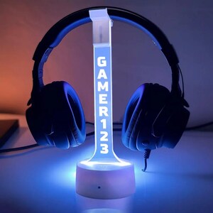 Puede incluir: Unos auriculares para juegos negros descansan sobre un soporte para auriculares iluminado en azul. El soporte muestra el texto "GAMER123" en blanco. La base y el soporte del soporte emiten un suave brillo azul, creando un aspecto moderno y elegante.