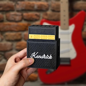 Könnte beinhalten: Ein schwarzer und gelber Miniaturverstärker mit dem Namen "Kendrick" in weißer Schrift. Der kleine Lautsprecher hat eine strukturierte Front und ist auf einem anderen schwarzen Lautsprecher gestapelt. Eine rote Gitarre ist im Hintergrund verschwommen.