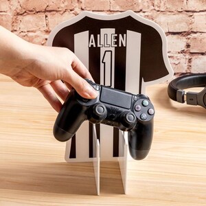 Puede incluir: Decoración en forma de camiseta en blanco y negro con el nombre "ALLEN" y el número "1" impresos. Un mando de videojuegos negro se sostiene frente a la camiseta. Unos auriculares negros son visibles en el fondo.