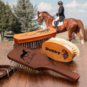 May include: Three wooden horse grooming brushes with personalized engravings. One brush is rectangular with the word "Faith" and a horse graphic. Another is oval with "Bobby" and a horse head. The third is paddle-shaped with "Bobby" and a unicorn.