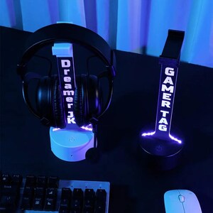 Puede incluir: Dos soportes iluminados para auriculares gaming. Uno muestra "Dreamer JK" en blanco, sosteniendo unos auriculares negros. El otro muestra "GAMERTAG" en blanco. Ambos soportes tienen un botón de encendido.